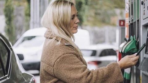 A woman with blonde hair wearing a brown coat pays for fuel at a pump in a petrol station