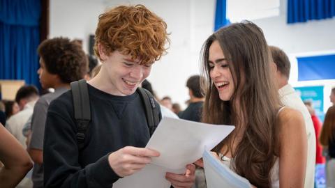 A young women and a young man in a crowded gymnasium looking down at a sheet of white paper the boy is holding. they both have broad smiles