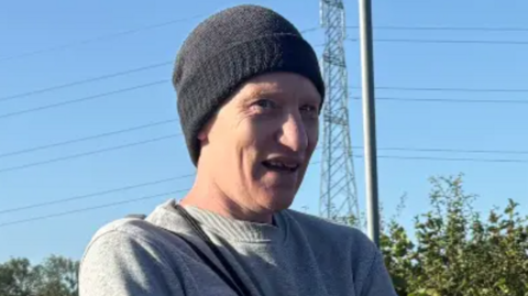 A man wearing a grey long-sleeved top and black beanie looks away from the camera. He is white.