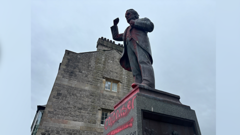 A statue of David Lloyd George with red graffiti saying 'coloniser' on the plinth and some spray paint on Lloyd George himself. There is a three-storey building behind the statue and the chimney is visible. It is a cloudy day.