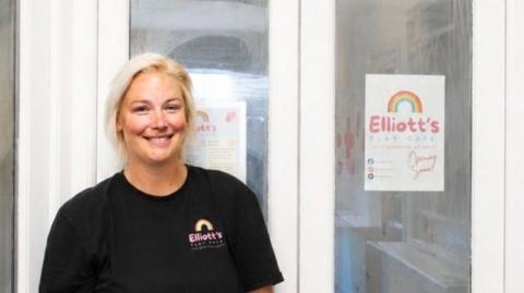 A person wearing a black Elliott’s Play Cafe T-shirt stands in front of white double doors beneath a large wooden sign that reads “Elliott’s Play Cafe – Little Adventures, Big Smiles.” Posters with the cafe logo are displayed on the glass panels of the doors. The interior features light-colored walls and pastel accents.