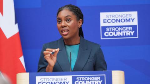 Badenoch is pictured from below the podium where she is giving a speech, at London Scottish House in Westminster, with a royal blue background and a Union flag to her left. She wears her braided hair tied back in a low ponytail, with a navy jacket and a turquoise top.