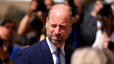 Prince William wears a navy blue suit and tie as he squints in the sunshine