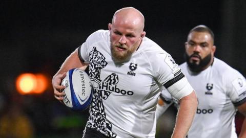 Josh Caulfield running with a rugby ball under his right arm held to his body during a match for Bristol in 2024. He is wearing a white Bristol kit of the 2024-25 season.