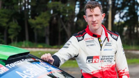 Elfyn Evans in a red and white jacket stands with one hand on his car windscreen in front of trees.