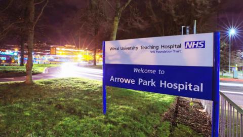 An external view of Arrowe Park Hospital, showing a blue and white sign on a grassy area. The glare of lights along a road coming out of the hospital can also be seen.