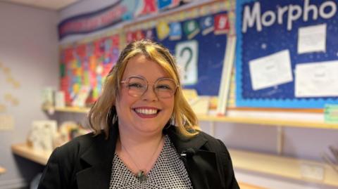 Katrina Crickett is wearing glasses and wearing a black jacket. She is sitting in a school classroom
