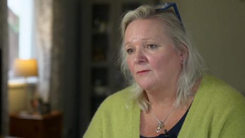 A woman in a light green cardigan and a black top is looking to the left of the camera. Her hair is just past her shoulders, she is wearing blue glasses that rest on the top of her head and a silver necklace with an anchor hanging from the chain. The background includes pale green walls a side table lamp with lampshade, a side table dark olive wardrobe doors, several blurred photos in frames and blurred patterned curtains.