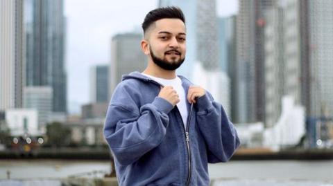 Kishan Thakrar, standing outside, holding his hooded top, standing in front of high rise buildings. He is wearing a white top, trousers and a blue hoodie, He is smiling at the camera and looking straight at the camera.