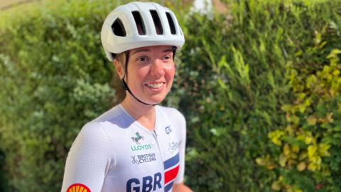 Lizzi Jordan wearing cycling kit with GBR on it and sponsor logos. She is also wearing a bicycle helmet and is smiling.