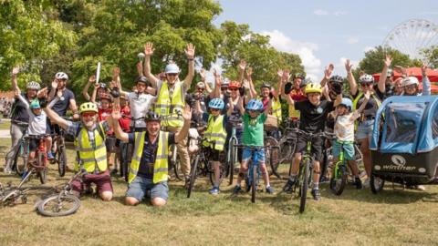 A group of around 30 men, women and children stand on a piece of grass in Stratford with trees behind them. They are stood or kneeling with their bikes and have bicycle helmets on. To the right of the image is a bike trailer for towing small children and in the far background is Stratford's ferris wheel.