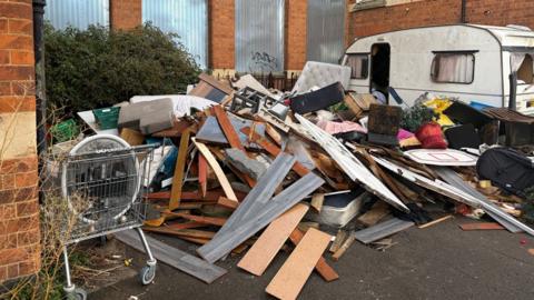 A large pile of dumped rubbish - mostly wooden flooring planks, broken pieces of wood from furniture, a grubby mattress, a supermarket trolley, and a caravan - which has been left outside a derelict brick building with corrugated windows.