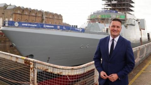 Streeting is smiling and standing in front of a war ship in dry dock, during a visit to a shipbuilding facility in Rosyth. He is wearing a navy suit with a white shirt and a black tie. He has brown, slicked-back hair.