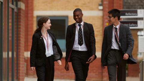 Three teenagers are walking side by side smiling at each other. On the left is a girl with dark curly hair in a ponytail. In the middle is a black boy. On the right is an Asian boy with glasses. They are all wearing black blazers, black trousers and white shirts. Both of the boys are wearing black ties.