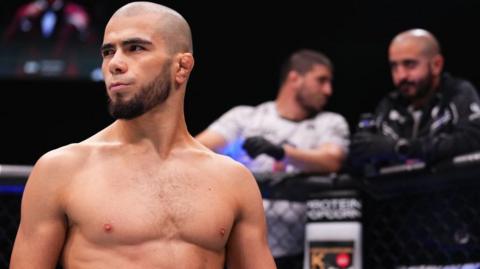 Muhammad Mokaev is shirtless and standing inside a mixed martial arts cage