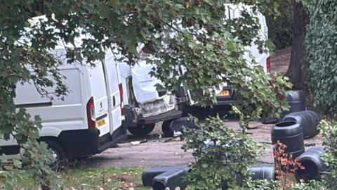Three white vans with a load of black barrels out the back. One of the vans has its doors blown off.
