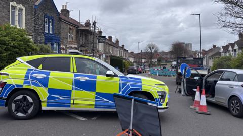 A police car with yellow and blue hi-vis decals, parked in the middle of Speedwell Road where a shooting has taken place. It is a cloudy overcast day and there are houses lining either side of the closed street.