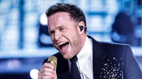 Olly Murs sings into a golden microphone, wearing a shirt, tie and blue suit.