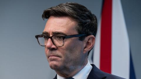 Britain's Prime Minister Andy Burnham attends a meeting with Canada's Prime Minister Mark Carney, he wears a black suit and white shirt. A Union flag hangs in the background.