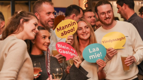 A group of people gathered together holding up speech bubble signs of Manx phrases.