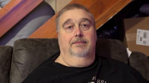 Image of a man with a white short beard, and short hair sitting in a leather chair wearing a black t-shirt. In the background can be seen some wooden stairs.
