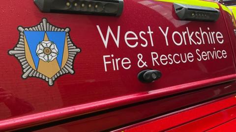 The side of a red fire engine, with white lettering that reads West Yorkshire Fire & Rescue Service, with a logo.