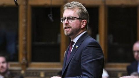 A man in a navy suit, purple tie and white shirt stands as he addresses the Houses of Commons. He has fair hair, a beard and dark-frame glasses. Other men are seen blurred on benches behind him in the wood panelled room.