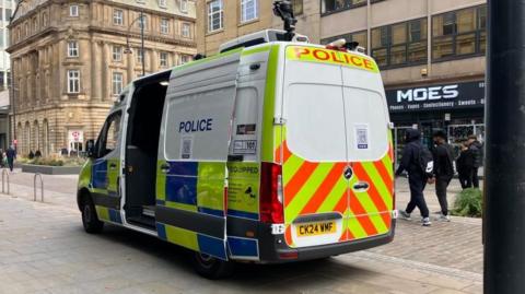 a police van is parked in a pedestrian area. There is a black camera mounted on the roof