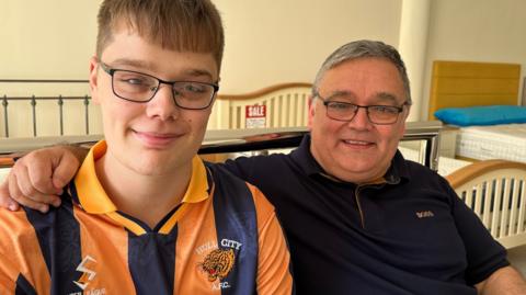 A father and son, both wearing glasses, sit together. The son is wearing a vintage amber and black Hull City shirt. His father is wearing a dark coloured polo shirt. He has his arm around his shoulder.