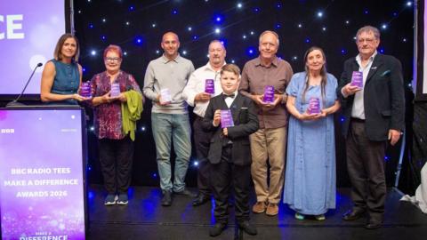 Eight award winners. From left to right: Catherine Devereux, a woman with brown bobbed hair, wearing a blue dress with buttons; Pauline Wilson, a woman with short red hair and wearing spectacles and a purple patterned top with a lime green shawl or cardigan in the crook of her left arm; Carl Trattles, a man with a bald or shaven head wearing a grey polo shirt and jeans; Ian Milburn, a balding man with a close cropped beard, wearing a white shirt; Roman, a young teenager with slicked back brown hair, wearing a black suit, white shirt and bow tie; Stuart White, an elderly man with grey hair, wearing a brown shirt and lighter brown trousers; Elizabeth Robinson, a middle-aged woman with long dark hair, wearing a blue dress and over-dress in the same colour; Ian Lawrence, a middle-aged man with short brown hair, wearing a dark jacket over a white shirt and dark trousers. All are holding their awards, which are rectangular purple objects with white lettering.