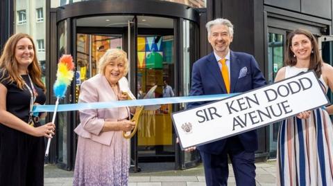 Kari Singleton, with long brown hair wearing a black dress holding a multi-coloured tickle stick, Lady Anne Dodd holding a giant pair of scissors about to cut a blue ribbon, with Spencer Phillips with grey hair and beard in a blue suit and orange tie and and Jill Carruthers with brown hair and wearing a white dress with blue and orange stripes holding a street sign which reads: "Sir Ken Dodd Avenue". They are all standing outside Showtown museum and are smiling.