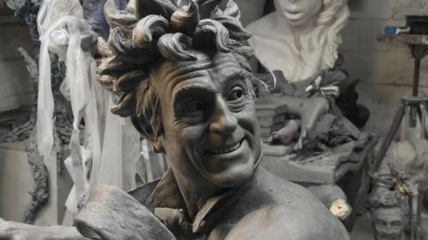 A close up picture of the clay model used for the sculpture showing Terry Jones with wild hair and smiling. He wears only a tie that looks like it is blowing in the wind