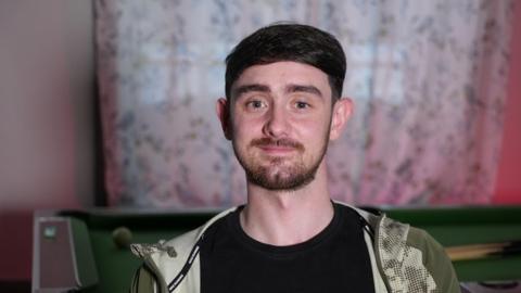 A head and shoulders picture of a man in his 20s looking straight into the camera and smiling slightly. He has short dark brown hair, dark prominent eyebrows and brown eyes. He has a short brown beard and moustache and is wearing a black t-shirt with an olive-green, patterned tracksuit-style jacket open over the top. A pool table is visible behind him, and some white patterned curtains are drawn across the window behind that.
