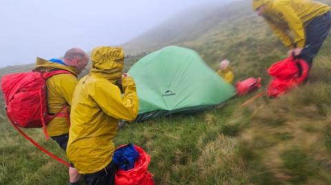 Skye Mountain Rescue Team - four men in yellow waterproof jackets and red rucksacks on a rainy hillside where there is a small green tent