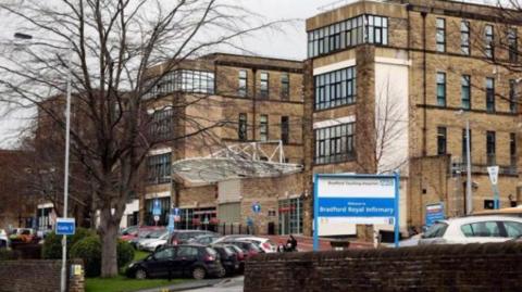 The outside of Bradford Royal infirmary, a large, multi-storey light brick building. Cars are parked outside the entrance.