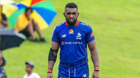 Joji Nasova pictured in the field for Fijian Drua while a lady shading under an umbrella on a grassy bank looks on