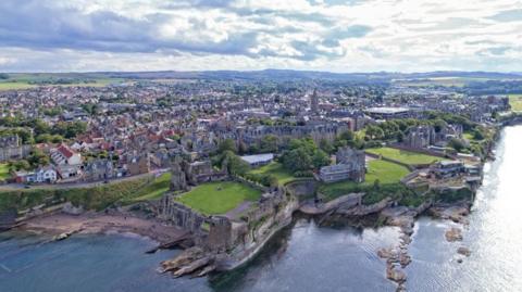 A birds eye view of St Andrews with a ruined cathedral next to the sea in the foreground, and the rest of the town in the background