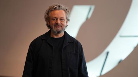 Michael Sheen who has curly, grey hair, a short beard and is wearing a dark shirt and jumper. He is smiling at the camera