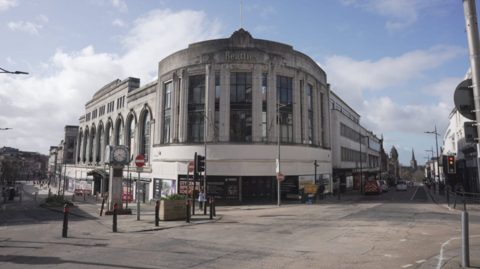 The photo shows the building standing on the corner of two streets in the city centre. It looks closed with a grubby concreate facade.