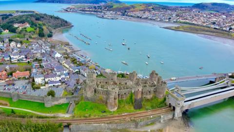 An aerial drone shot of the railway in Conwy. It passes by Conwy Castle, with the sea beyond.