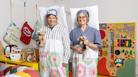 Two women dressed in night dresses and their bed costumes, holding teddy bears in a children's play room.