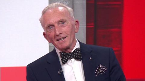 A man with grey hair and blue eyes wears a navy blue suit jacket, a white shirt, and a dark spotted bow tie with a matching pocket square. He sits in front of a red studio background.