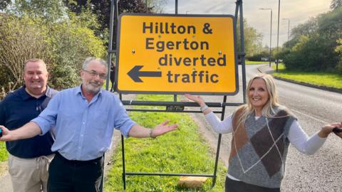Two men and a woman stand by a yellow temporary road sign. The sign reads “Hillton & Egerton diverted traffic.”