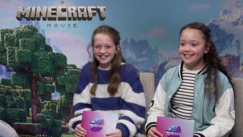 Two girls smiling in front of a Minecraft Movie backdrop, they are holding Newsround cue cards.