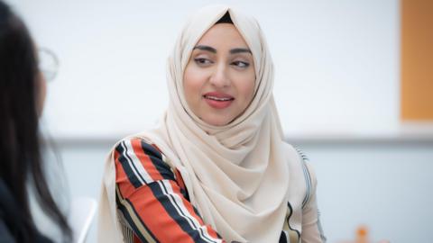 Syeda Batool sitting down, looking at a person to her left. She has a scarf covering her head, and is wearing a striped long sleeved top.