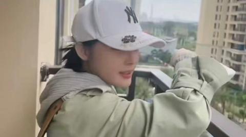 A woman is standing on the balcony of a high rise building, overlooking other apartment blocks and some green trees on the ground. There is a cityscape behind. The woman has dark hair and is wearing a white New York Yankees baseball cap. She has a light coloured hooded jumper, and a green mackintosh style coat over the top.