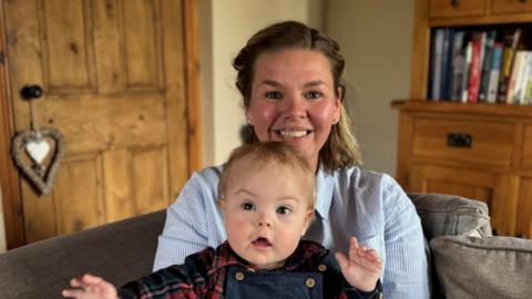 Emma with her son Bert, who's holding his hands up. She is smiling and has her hair tied back, wearing a blue and white striped shirt.