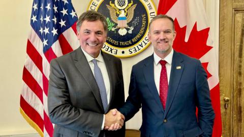 An image showing US trade representative Jamieson Greer (right) shaking hands with Canada-US trade minister Dominic LeBlanc to his left. Behind them are a US and Canada flag with the seal for the US trade representative in the middle. Both men are wearing suits and smiling at the camera.