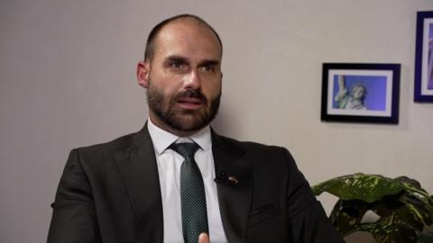 Eduardo Bolsonaro is speaking as he sits against a white, blank background with a plant and a close-up photo of the Statue of Liberty visible to his left. He is in a dark suit, white shirt and dark patterned tie.