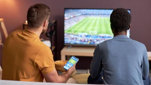 Two men watching football on a TV with one holding a mobile phone with a betting site showing